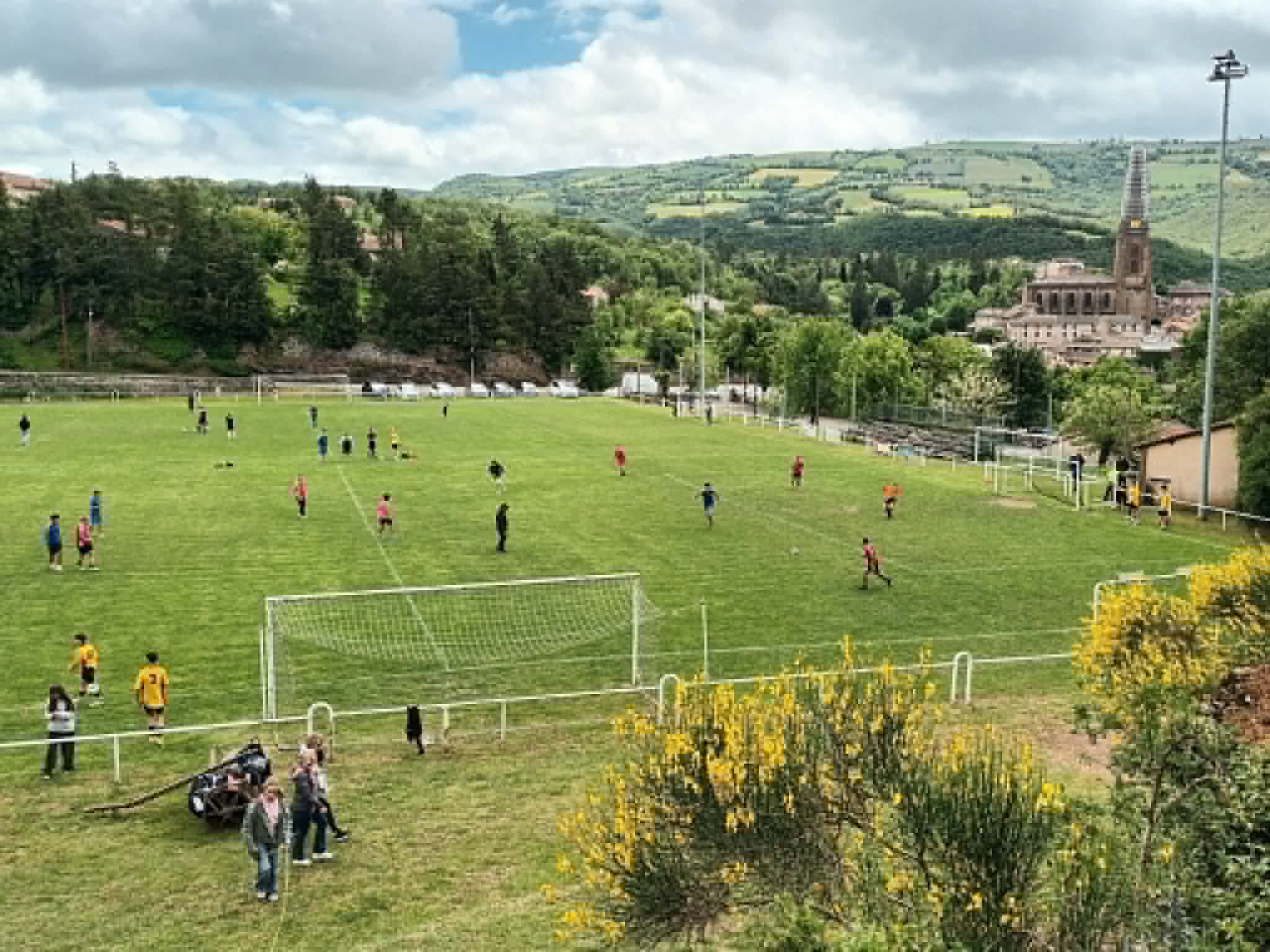 Vue du stade municipal de Belmont-sur-Rance pendant le tournoi de sixte de l’Ascension : des équipes jouent sur la pelouse, avec le clocher de l’église et les collines en arrière-plan.